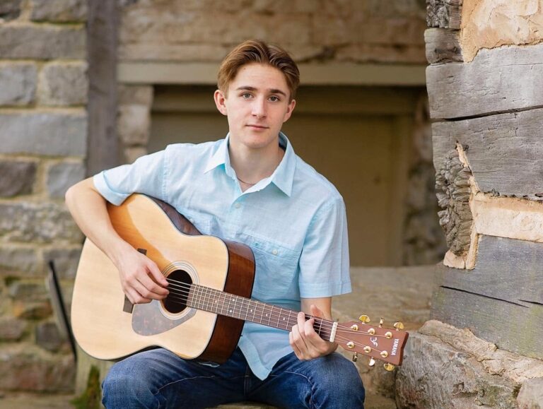 Caleb Ronne sitting in front of a building, holding a guitar. He is wearing a light blue dress short and blue jeans. In this entry, Jess Ronne write about her parenting journey with Caleb, and asks him hard questions about what it was like growing up with a special needs sibling, Lucas.