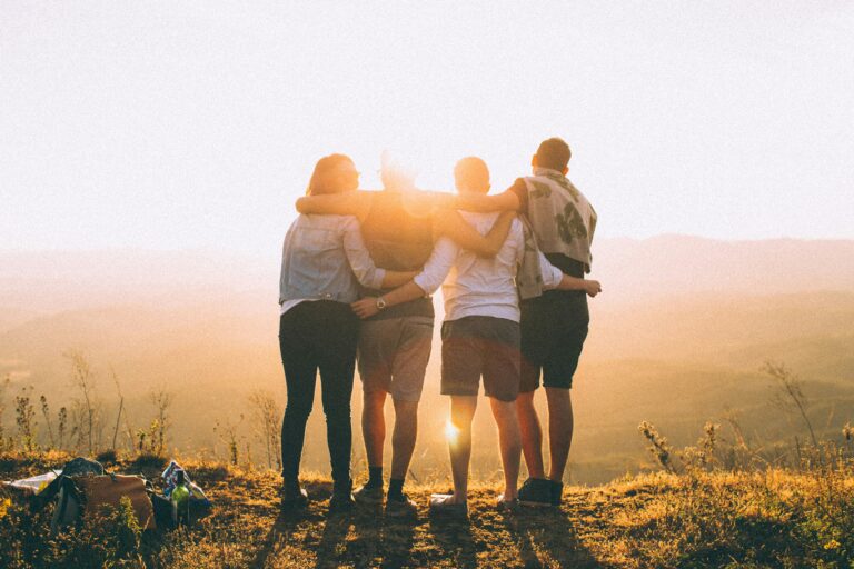 Group of people standing in a field, looking at the sunset, their backs are to us. Everyone is locking arms. This is our tribe. Photo by Helena Lopes on Unsplash