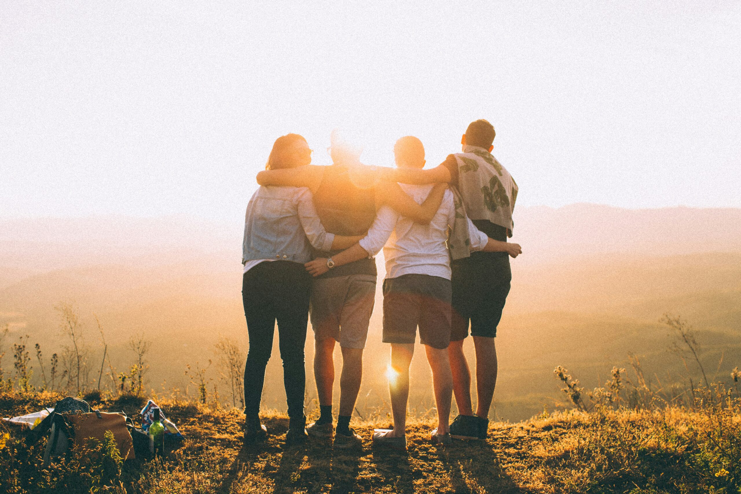 Group of people standing in a field, looking at the sunset, their backs are to us. Everyone is locking arms. This is our tribe. Photo by Helena Lopes on Unsplash